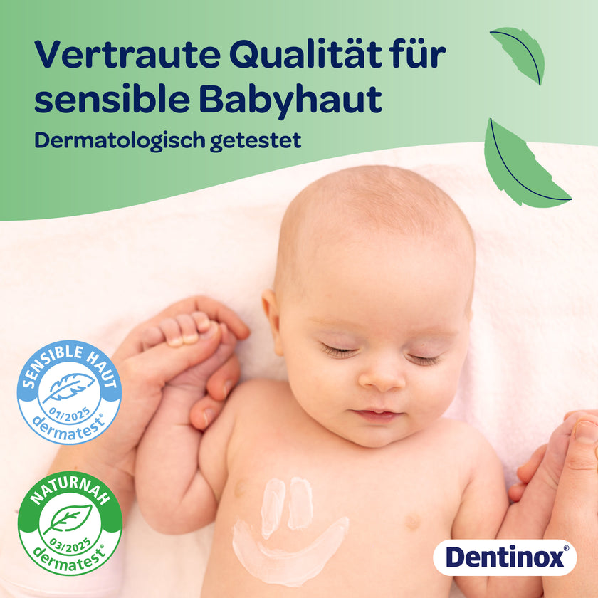 Baby mit Creme-Smiley auf der Brust und Händchen haltend mit einer Erwachsenen Person auf Decke, Schrift und Siegel im Vordergrund