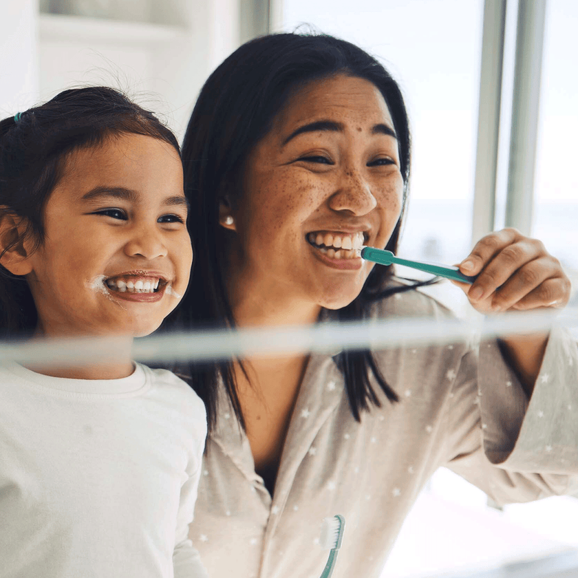 Mutter und Tochter putzen sich die Zähne und lächeln dabei in den Spiegel